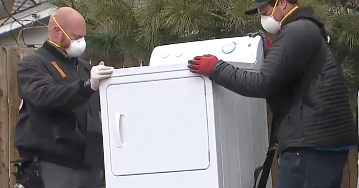 Two men wearing face masks and gloves moving a white dryer outdoors during delivery